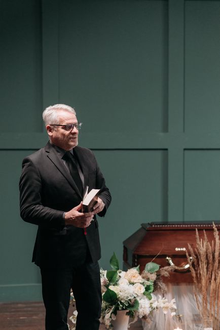 A man with gray hair and glasses, dressed in a black suit, stands indoors holding a small open book. Behind him, a teal wall provides a neutral backdrop. To his right, there is a floral arrangement fe
