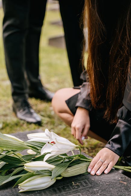 A close-up of a person kneeling on the ground, arranging a bouquet of fresh white lilies with green leaves. The lilies have large, trumpet-shaped petals and prominent stamens, appearing vibrant and re