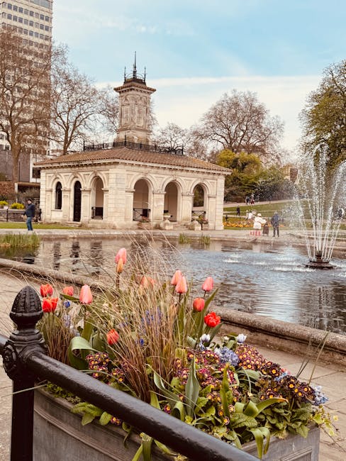 A vibrant floral arrangement placed in a large rectangular planter near a pond in a park. The bouquet includes pink tulips, red roses, purple pansies, and other mixed flowers with green foliage, all f