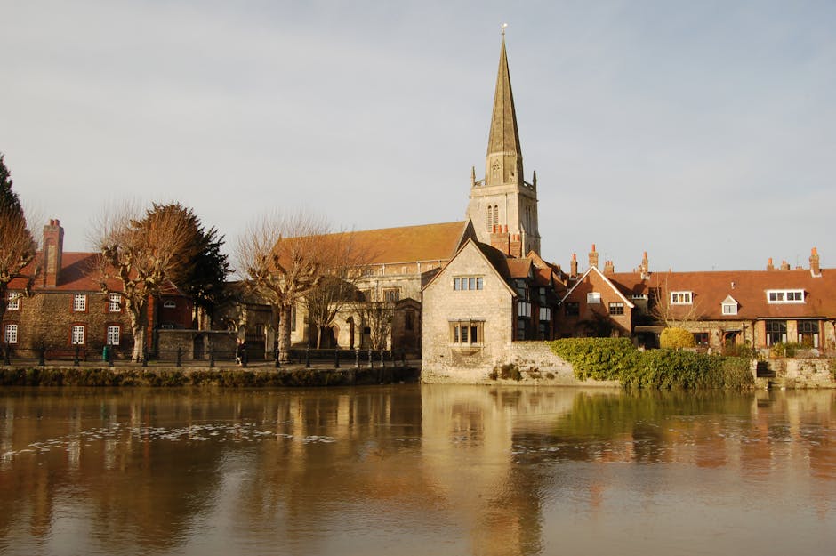 A riverside view featuring a historic church with a tall, pointed spire and a cluster of traditional red-brick houses with tiled roofs along the water's edge. The scene is illuminated by soft, natural