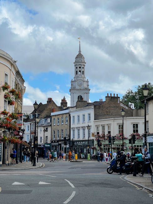 The image depicts a lively town square in Carshalton, with a prominent historic clock tower topped with a golden weather vane against a partly cloudy sky. Surrounding the square are multi-story buildi