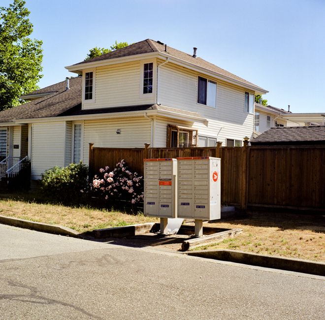 A close-up view of a residential street showing a white, two-storey house with a dark tiled roof, surrounded by a wooden fence and lush green trees. In front of the house, there's a small garden with