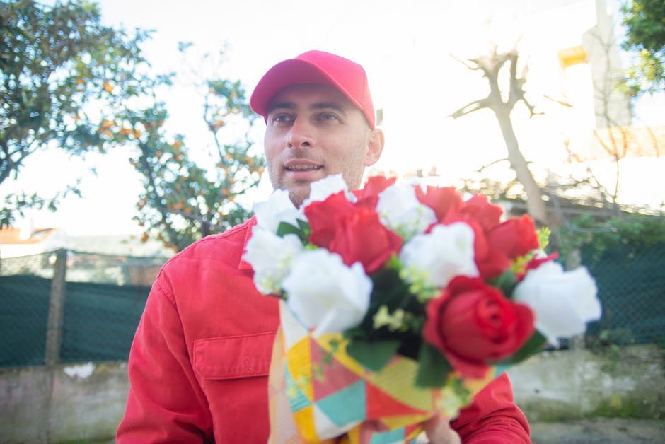 A male florist wearing a red cap and matching red jacket holding a vibrant floral bouquet outdoors during daylight. The bouquet features red and white roses, with lush green foliage, wrapped in colorf