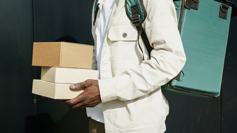 Close-up of a person holding three stacked rectangular boxes, two white and one brown, in their hand. The individual is wearing a cream-colored jacket and has a large green backpack slung over their s